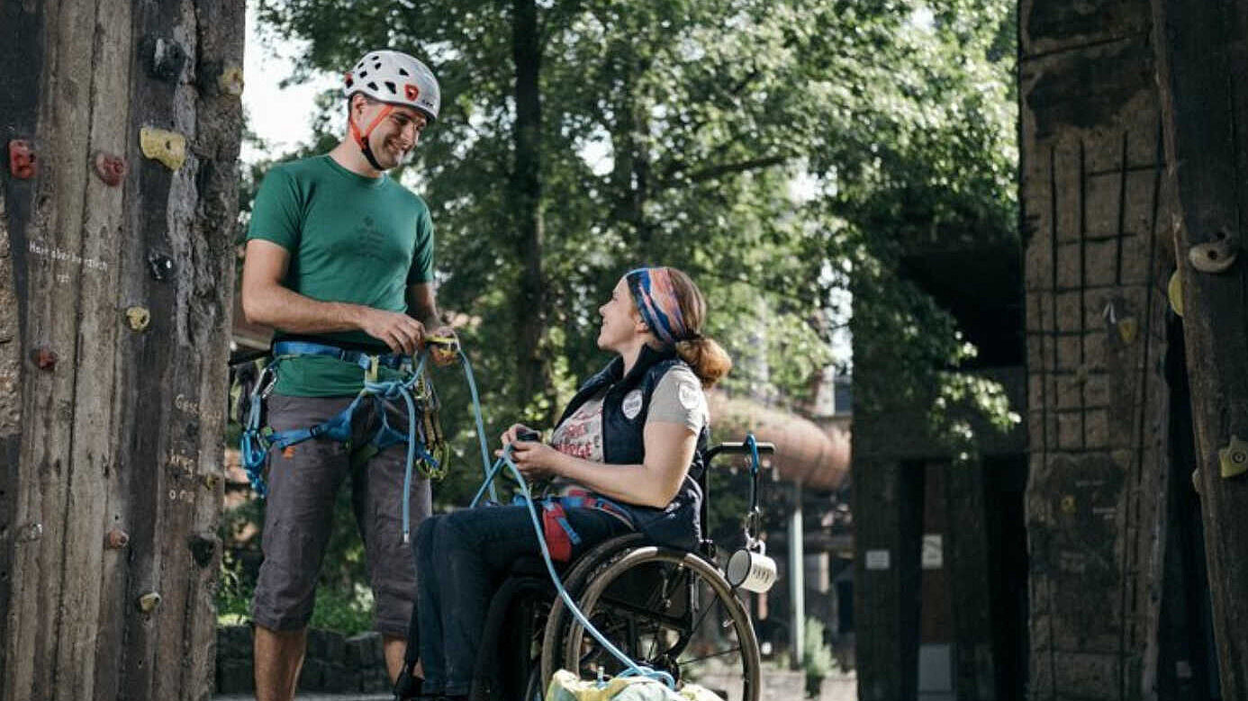 Ein Mann mit Helm und eine Frau im Rollstuhl vor einer Kletterwand.